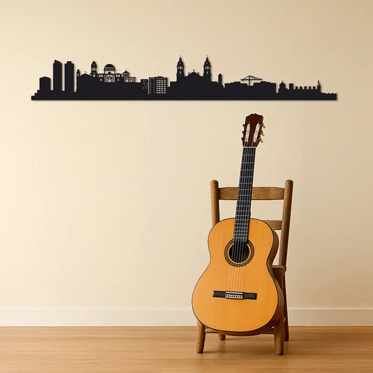 Skyline de Cádiz en madera DM sobre pared beige, acompañado de guitarra española apoyada en silla rústica. Decoración con esencia andaluza para amantes del flamenco, la música y la artesanía. Silueta de Cádiz ideal para hogares con alma del sur.