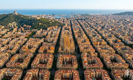 Vista aérea de la ciudad de Barcelona, destacando una gran catedral de estilo gótico en el centro, rodeada de edificios residenciales con tejados rojizos y calles arboladas; al fondo se aprecia el mar y una colina con vegetación.