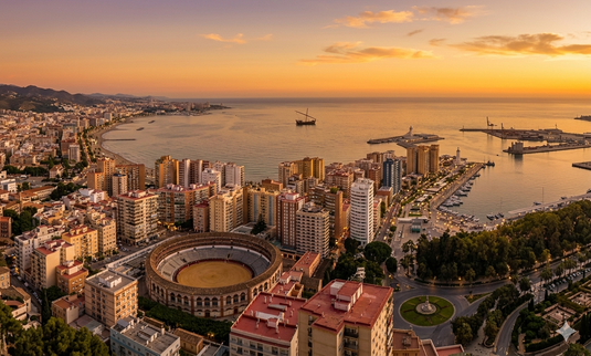 Vista panorámica de Málaga desde las alturas, con la Plaza de Toros de La Malagueta en primer plano, edificios residenciales, el puerto deportivo y el mar Mediterráneo extendiéndose hacia el horizonte bajo un cielo despejado.